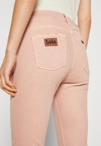 Vaaleanpunaiset farkut, joissa on istuva muotoilu. Hihassa on ruskea nahkapatri, jossa on "Lois"-brändäys ja kaksi takataskua. Pehmeä kangas.