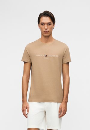 Giovane uomo che indossa una t-shirt beige Tommy Hilfiger con logo e pantaloncini bianchi, in piedi davanti a uno sfondo chiaro e uniforme.