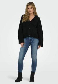 Cardigan nero lavorato a maglia con scollatura a V, chiusura con bottoni frontali e due tasche a toppa, abbinato a jeans blu in denim e stivaletti neri.