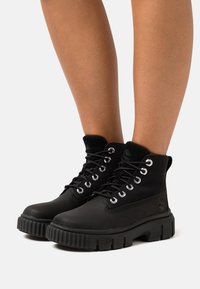 Bottines noires avec un extérieur en cuir lisse, semelle en caoutchouc robuste, œillets en métal et doublure en textile. Présente un logo sur le côté.