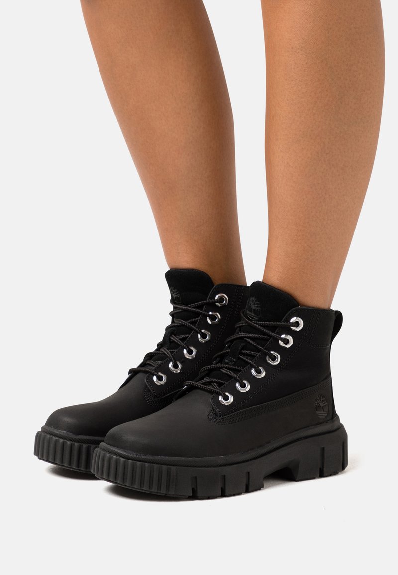 Bottines noires avec un extérieur en cuir lisse, semelle en caoutchouc robuste, œillets en métal et doublure en textile. Présente un logo sur le côté.