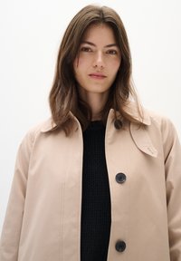 Beige deštník s límcem, se černými knoflíky a hladkou, voděodolnou texturou. Nesený přes tmavý pletený svetr.