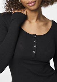 Svart ribbad långärmad topp med rundad halsringning och fem tryckknappar på bröstet. Tyget har en jämn textur med en åtsittande design.