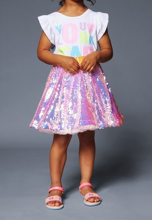 Enfant portant une chemise blanche à manches volantées avec des lettres colorées et une jupe rose à sequins, debout les mains jointes et des sandales roses.