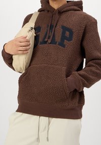 Hnědá fleecová mikina s kapucí s logem GAP, elastickými manžetami a přední kapsou, doplněná béžovou kabelkou přes rameno. Texturovaná, teplá látka.