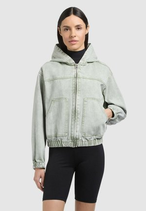 Femeie purtând o jachetă zip-up cu glugă, de culoare gri deschis, cu buzunare mari în față și manșete elastice, asortată cu pantaloni scurți negri pentru ciclism, cu aspect nervurat.