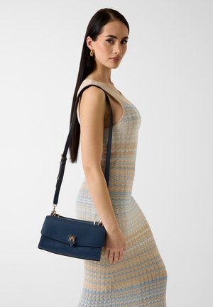 Frau mit langen dunklen Haaren, die ein ärmelloses, gemustertes Kleid trägt und eine dunkelblaue Umhängetasche mit goldenen Beschlägen bei sich hat.