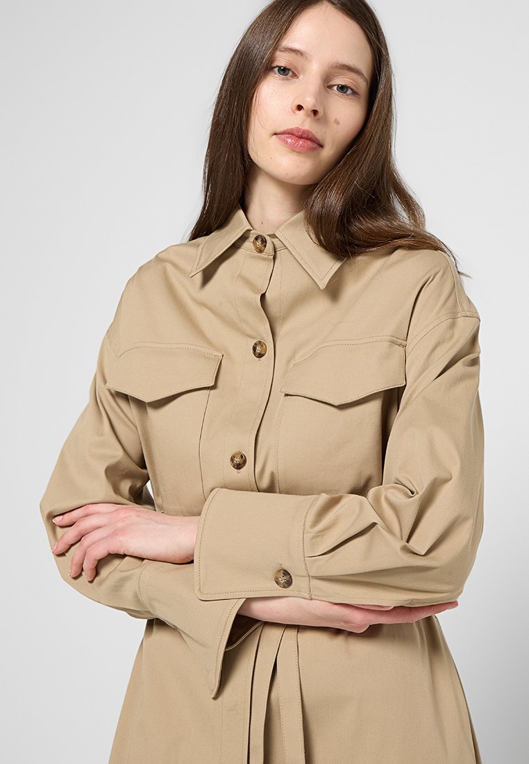 Beżowa koszula zapinana na guziki z dwoma kieszeniami na piersi, długimi rękawami i dopasowanym krojem. Posiada duże guziki oraz gładką teksturę materiału.