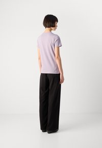 Moteris su trumpais plaukais, vilkinti šviesiai violetinę trumpomis rankovėmis marškinėlį ir juodas plačias kelnes, stovi ir žiūri į šalį priešais vientisą baltą foną.