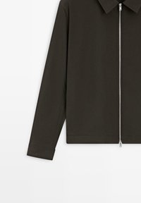 Donkergroene lange mouwen jas met een kraag en een zilveren ritssluiting aan de voorkant, gedeeltelijk zichtbaar tegen een witte achtergrond.