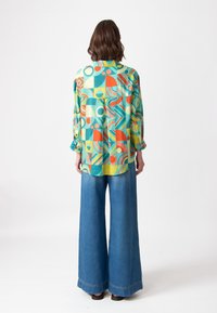 Camisa de botones colorida con patrones geométricos en teal, naranjas y amarillos, combinada con pantalones anchos de mezclilla azul de corte holgado.