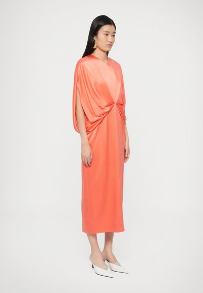 Κοραλλί midi φόρεμα με φουσκωτή σιλουέτα, διαθέτοντας διπλωμένη εμπρόσθια λεπτομέρεια και φαρδιά μανίκια. Λευκά mules με μυτερή μύτη ολοκληρώνουν την εμφάνιση.