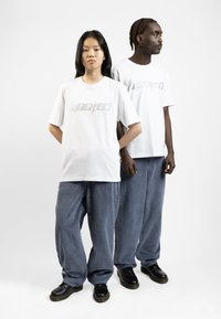 T-shirt bianchi con la scritta "Wasted" stampata in un finish lucido, abbinate a pantaloni larghi in corduroy blu e calzature nere.