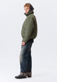 Olivgrüne Steppjacke mit Kapuze, kombiniert mit dunkelblauen Denim-Jeans und schwarzen Stiefeln mit grauen Akzenten. Glatte Textur und lässiges Design.