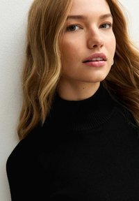 Giovane donna con capelli biondi lunghi, che indossa un dolcevita nero, guardando leggermente di lato contro uno sfondo bianco.