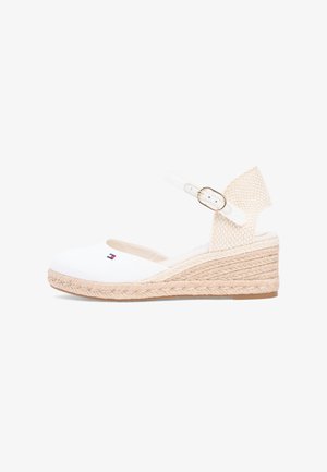 Witte canvas sleehaksandaal met beige gevlochten zool, verstelbare enkelriem met gesp en een klein rood-wit-blauw logo aan de zijkant.
