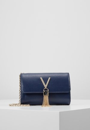 Laivaston sininen teksturoitu clutch-laukku kultaisen V-muotoisen lukon, tupsuketjujen ja vastaavan kultaisen ketjuhihnan kanssa valkoisella jalustalla.