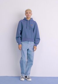 Haut à capuche violet clair avec poche kangourou, associé à un jean bleu clair et des baskets noir et blanc avec un motif rayé.
