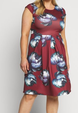 Kvinde iført en ærmeløs, vinrød kjole med store blå og hvide blomsterprint, stående foran en ensfarvet lysegrå baggrund.