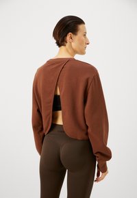 Langärmliger brauner,cropped Sweatshirt mit einem offenen Rückendesign. Kombiniert mit hoch geschnittenen braunen Leggings, beides aus weichem, texturiertem Material.