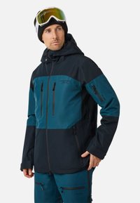 Man in een teal en zwart skijack en -broek, zwarte beanie en witte skigoggles, die iets naar links kijkt met zijn hand in zijn zak.