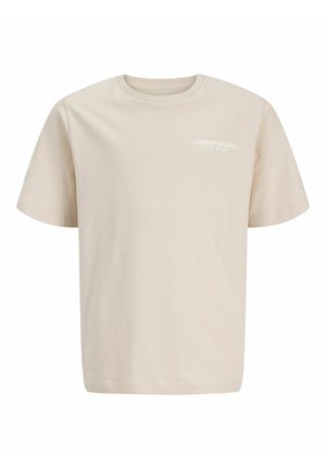 Garçon debout, portant une chemise à manches courtes noire et blanche à motif floral, un t-shirt beige, un jean bleu et des baskets blanches, regardant vers sa droite.