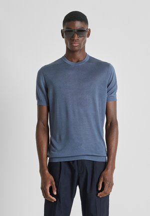 Man in blauw shirt met korte mouwen en donkere broek, draagend grote zwarte zonnebril, staand tegen een effen grijze achtergrond.