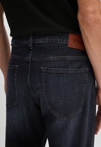 Jeans in denim blu scuro con una texture liscia, dotati di due tasche posteriori e un'etichetta in pelle marrone con la scritta "BOSS" sulla vita.