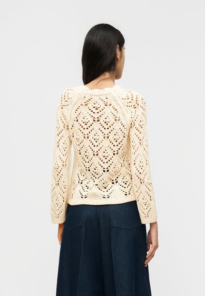 Γυναίκα με μακριά σκούρα μαλλιά που φοράει ένα κρεμ χρώματος πλεκτό πουλόβερ με μακριά μανίκια και σκούρο μπλε ψηλόμεσο παντελόνι, γυρισμένη προς τα πίσω.