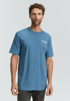 Camiseta de algodón azul con mangas cortas y cuello redondo. Presenta un logo blanco impreso en la parte izquierda del pecho. Diseño simple y sólido.