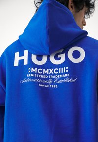 Blå hettegenser med en stor hvit "HUGO" logo, intrikate tekstdetaljer og snor i hetten. Myk tekstur er synlig.