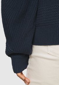 Granatowy dziany sweter z fakturowanym wzorem i ściągaczami przy rękawach. Zbliżenie na rękaw ukazuje szczegółowy splot i elastyczny materiał.