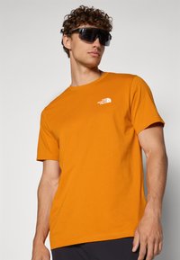 Tricou scurt cu mânecă, de culoare portocalie, fabricat din bumbac, cu guler rotund. Are un logo alb pe piept. Modelul poartă ochelari de soare supradimensionați, negri.