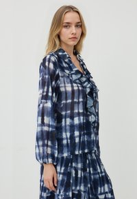 Blusa a maniche lunghe blu e bianca con colletto arricciato, realizzata in un tessuto liscio. Presenta un motivo a scacchi con sfumature.