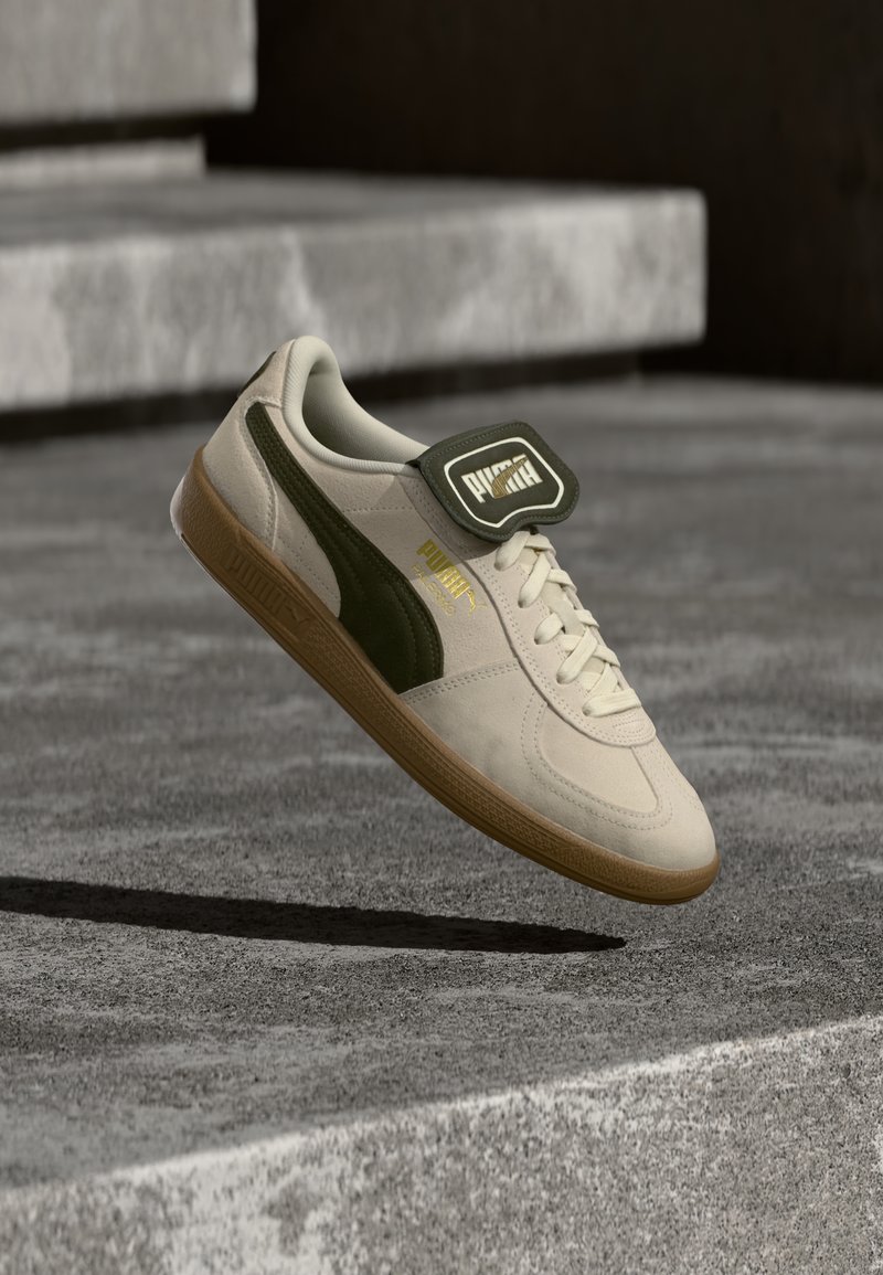 Sapatilha de camurça bege com detalhes em verde escuro, sola de borracha, língua acolchoada e detalhe em dourado na marca. Flutuando acima de uma superfície cinza texturizada.