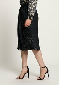 Donna che indossa una gonna nera plissettata, una blusa a stampa leopardata e sandali neri con tacco alto e cinturino alla caviglia, in piedi su uno sfondo chiaro.