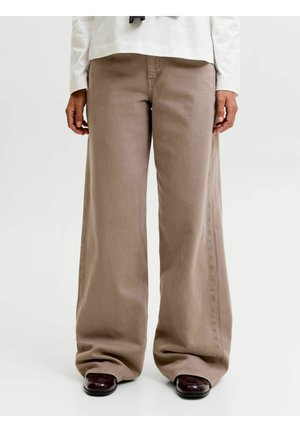 Persona che indossa pantaloni beige a gamba larga abbinati a scarpe marrone scuro con punta quadrata, in piedi su uno sfondo bianco uniforme.