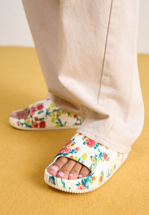 Pieds dans un pantalon beige portant des sandales blanches avec des motifs floraux colorés sur un sol en moquette jaune.