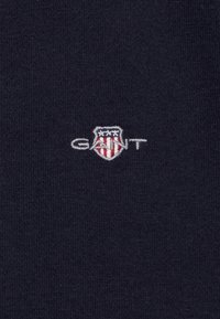 Tela de algodón azul marino con una superficie texturizada. Presenta un logotipo de escudo bordado en blanco y rojo con el texto "GANT".