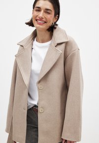 Beeži villasegu mantel suurte krae ja kahe esipaknaga, laiad varrukad, valge särke ja hallide pükstega. Sile tekstuur.