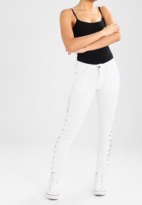 Jeans skinny blancos con detalles de cordones laterales, combinados con una camiseta ajustada negra y zapatillas altas blancas. Textura suave, material elástico.