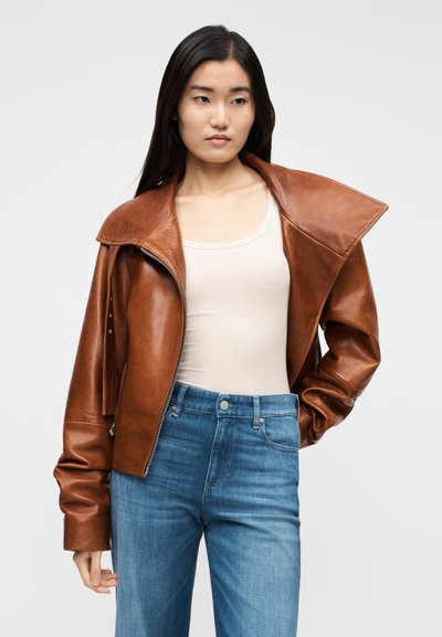 Giacca di pelle marrone con colletto ampio, chiusura con zip e spalle strutturate. Abbinata a jeans azzurri a vita alta. Texture liscia.