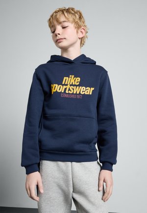 Marineblaue Kapuzenjacke aus weichem Stoff mit einem großen gelben Textlogo "nike sportswear" und einer Kängurutasche. Kombiniert mit grauen Jogginghosen.