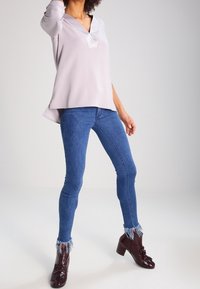 Blusa viola chiaro a maniche lunghe con scollo a V, abbinata a jeans skinny blu con orli sfrangiati e stivali con tacco borgogna.