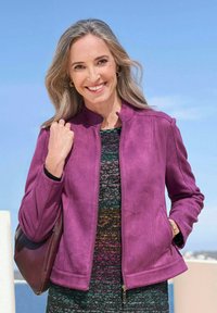 Veste en daim violette avec un col montant, fermeture éclair et poches latérales, associée à une robe à motifs texturés, portée avec un sac à main.
