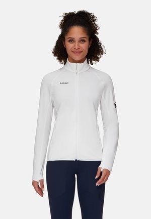 Donna con capelli ricci che indossa una giacca bianca con zip e pantaloni scuri, rivolta frontalmente con uno sfondo grigio chiaro neutro.