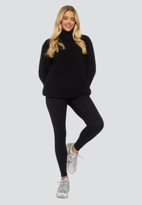 Pullover in pile nero con colletto alto e collo con zip; abbinato a leggings neri e sneaker argentate. Texture liscia con una finitura soffice.