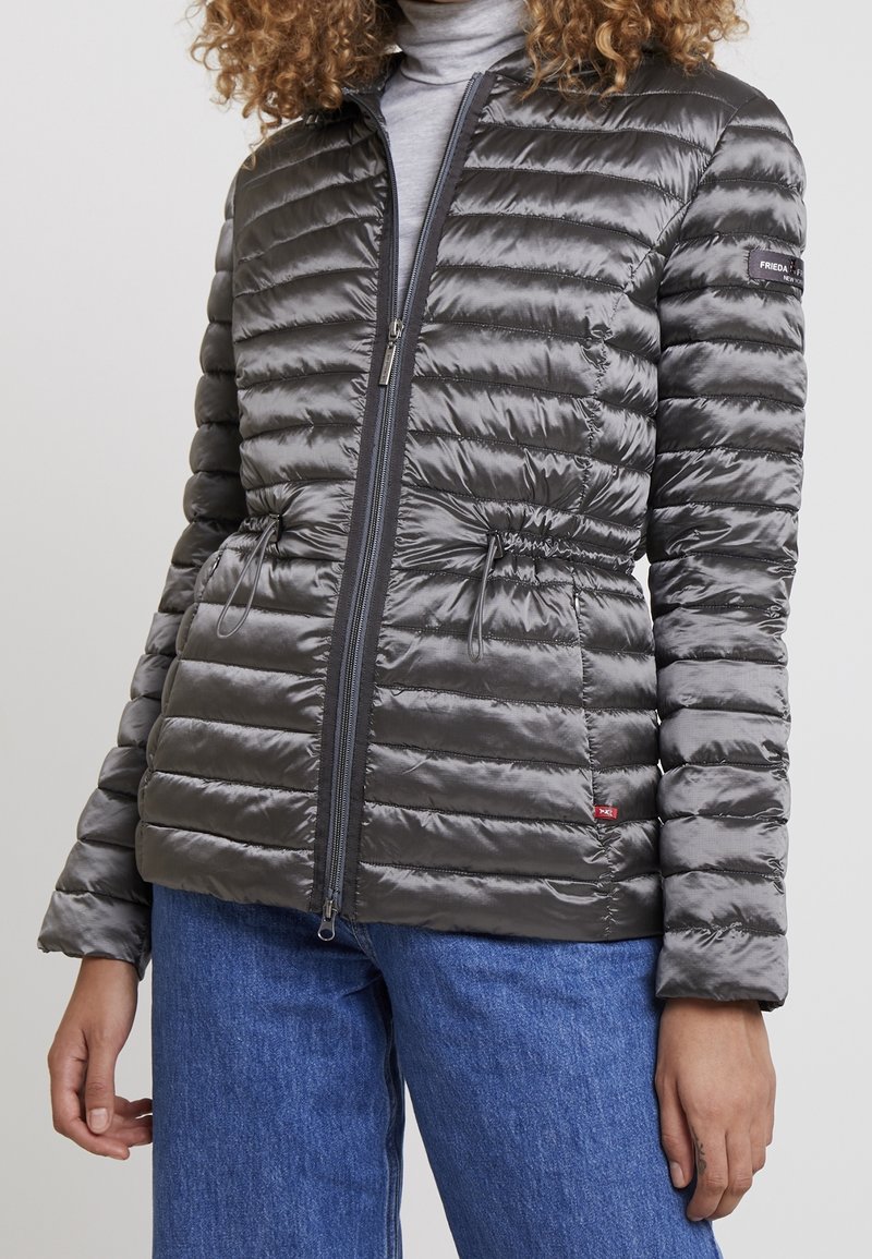 Graue Steppjacke mit gepolsterter Textur, Reißverschluss vorne, verstellbarem Kordelzug am Bund und Stehkragen. Kombiniert mit blauen Jeans.