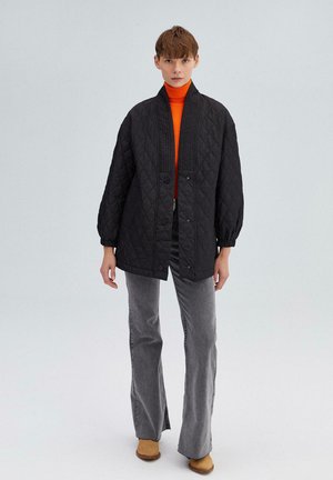 Black quilted jacket with a high collar, snap buttons, and elastic cuffs, worn over an orange turtleneck and paired with gray flared jeans.