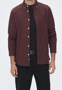 Bordeauxrotes Hemd mit Knöpfen aus strukturiertem Stoff, mit einer Brusttasche, weißen Knöpfen und langen Ärmeln, kombiniert mit einem schwarzen T-Shirt und einer Hose.